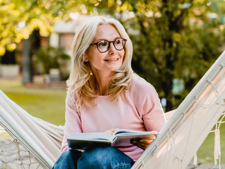 Entspannt und lächelnd liest eine blondhaarige Frau mit Brille in einer Hängematte ihr Buch in ihrem Garten.