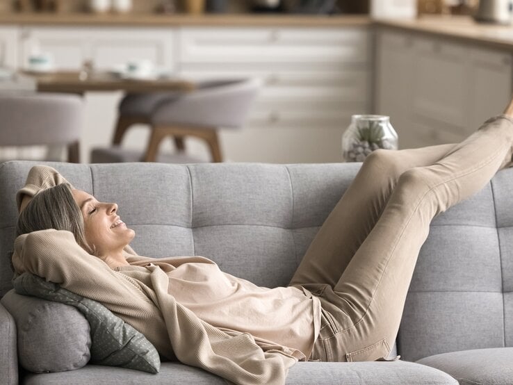 Gelassene Frau mittleren Alters mit geschlossenen Augen genießt entspannt den Moment auf einem Sofa in gemütlicher Umgebung.