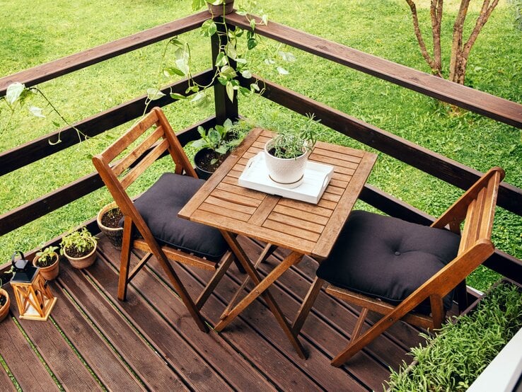 Ameisen auf Gartenmöbeln Zwei Holzklappstühle mit schwarzen Polstern stehen an einem kleinen Holztisch auf einer Terrasse mit Kräutertöpfen und Grünpflanzen.