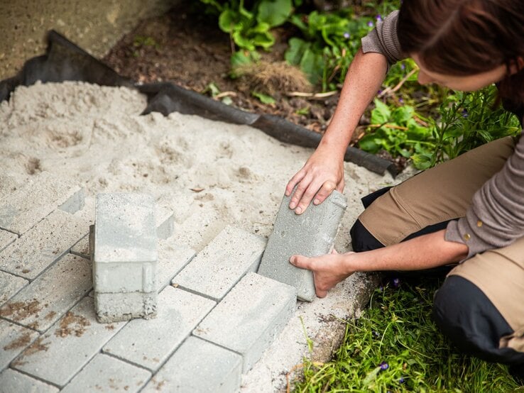 Pflastersteine richtige verlegen (Schichten) | © Shutterstock/Miriam Doerr Martin Frommherz Junge Frau mit braunen Haaren und Outdoorhose setzt kniend einen grauen Pflasterstein in ein Sandbett am Rand eines Gartens ein. | © Shutterstock/Miriam Doerr Martin Frommherz