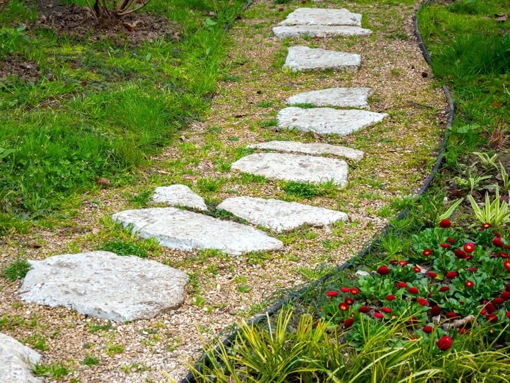 Unregelmäßig geformte Trittsteine führen geschwungen über einen Kiesweg durch einen gepflegten Garten mit frischem Grün.