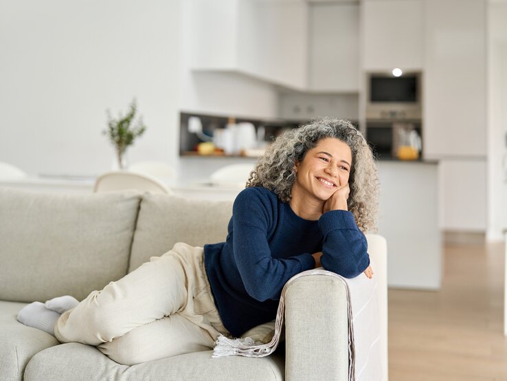 Eine grauhaarige Frau mittleren Alters liegt entspannt lächelnd in heller Jogginghose und dunkelblauem Pullover auf einem Sofa.