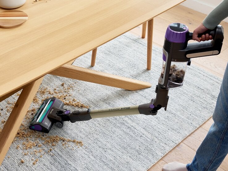 © SharkNinja Moderne Handstaubsaugerdüse mit violetten Akzenten entfernt Krümel unter einem Tisch auf hellem Teppich in wohnlicher Umgebung. | © SharkNinja