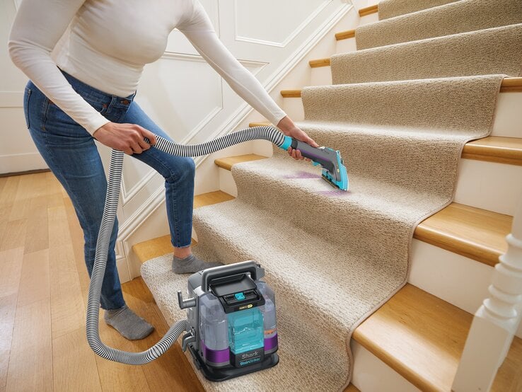 © SharkNinja Junge Frau in weißem Langarmshirt und Jeans reinigt konzentriert mit Teppichreiniger einen lilafarbenen Fleck auf beiger Treppe. | © SharkNinja