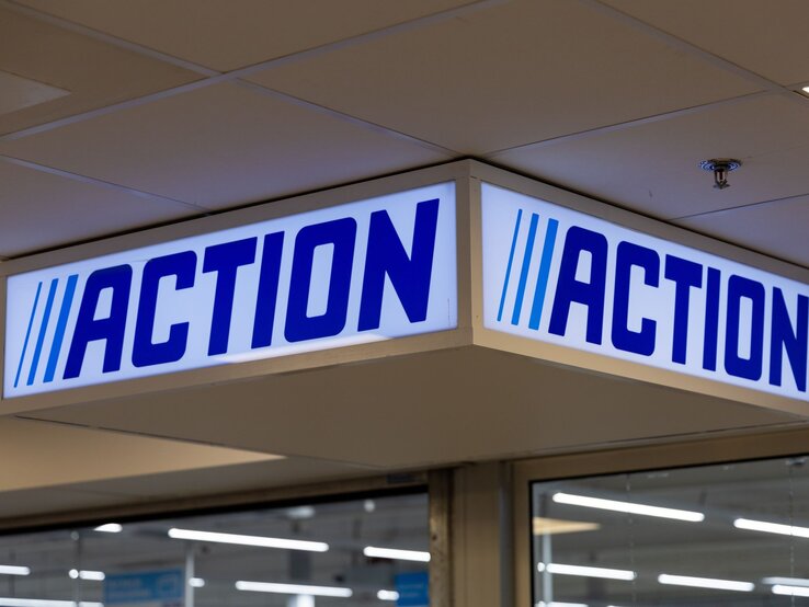 Leuchtendes Aktions-Schild mit blauem Schriftzug „ACTION“ und drei Streifen hängt unter weißer Decke im Eingangsbereich.