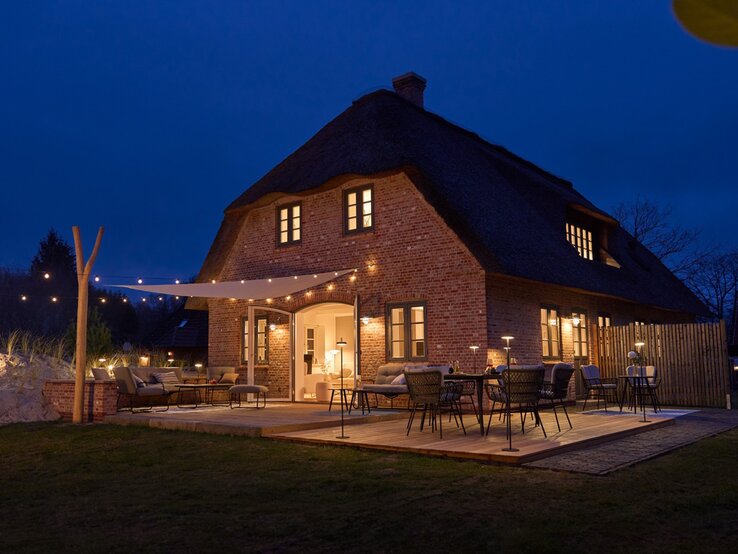 Gemütliches Reetdachhaus aus rotem Backstein bei Nacht, mit stimmungsvoll beleuchteter Holzterrasse und stilvollen Gartenmöbeln.