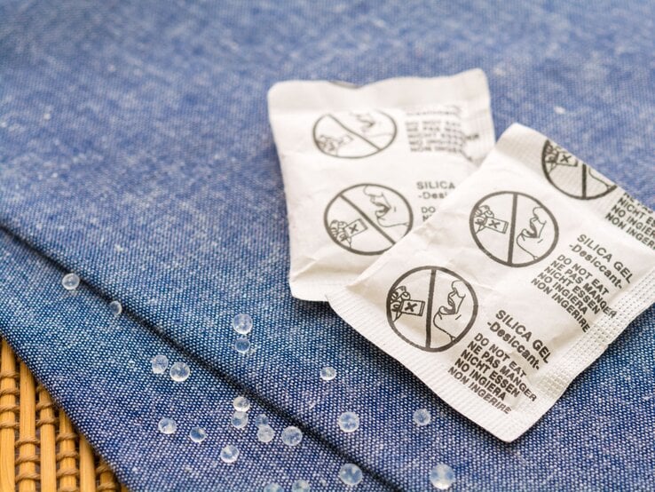 Zwei geöffnete Silicagel-Beutel mit Warnhinweis liegen auf blauem Stoff, daneben verstreute durchsichtige Kügelchen auf Leinenstruktur.