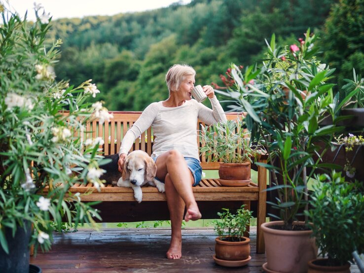 Ältere Frau mit kurzem grauem Haar sitzt barfuß auf Holzbank, streichelt schlafenden Hund und trinkt Kaffee im Garten.
