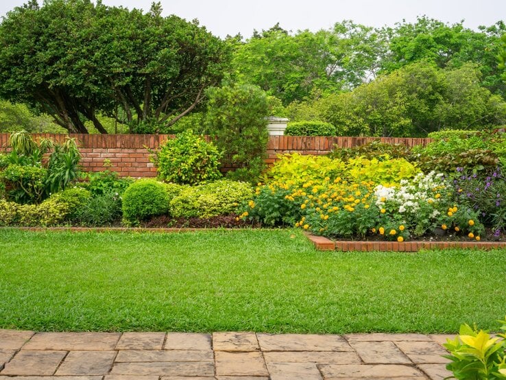 Gepflegter Garten mit sattgrünem Rasen, blühenden Beeten in Gelb, Weiß und Violett vor roter Backsteinmauer.