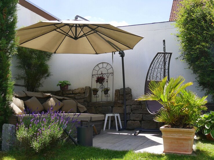 Garten Lounge kleiner Balkon Gemütliche Gartenecke mit Sonnenliege, Hängesessel, Sonnenschirm und blühendem Lavendel vor heller Mauer und Steinwand.