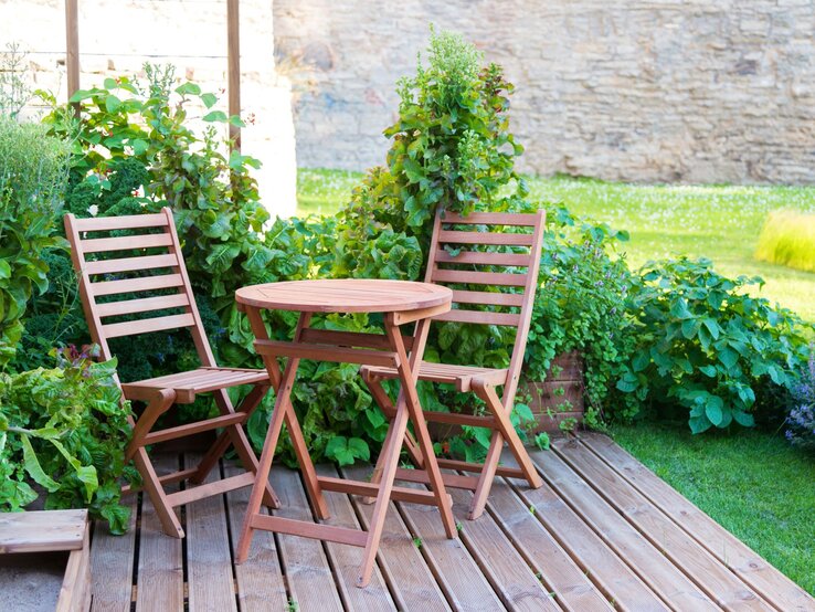 Garten Tisch Lidl Kleiner Holztisch mit zwei passenden Stühlen steht auf einer Holzterrasse, umgeben von üppigem Gemüse und Kräutern.