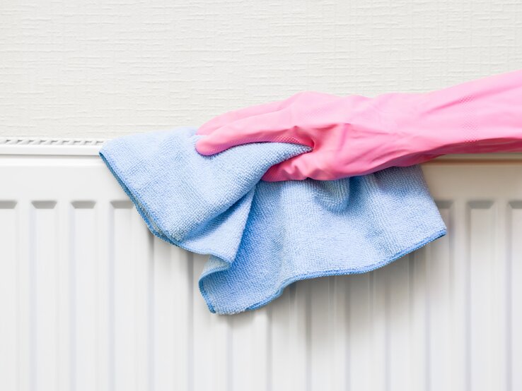 Heizung reinigen Eine Hand in pinkem Gummihandschuh wischt mit einem blauen Mikrofasertuch einen weißen Heizkörper vor cremefarbener Wand ab.