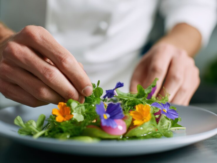 Ein Koch in weißer Kleidung garniert mit präziser Handbewegung einen frischen Salat mit essbaren Blüten und Kräutern auf einem Teller.