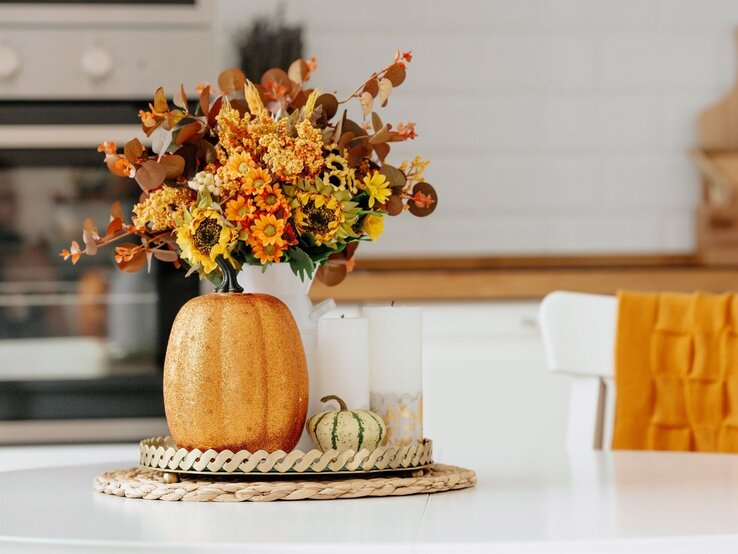 Herbstdeko aussortieren: Blumen | © Shutterstock/PhotoJuli86 Ein goldener Deko-Kürbis, ein herbstlicher Blumenstrauß in warmen Farben und Kerzen stehen auf einem Tisch vor heller Küchenzeile. | © Shutterstock/PhotoJuli86