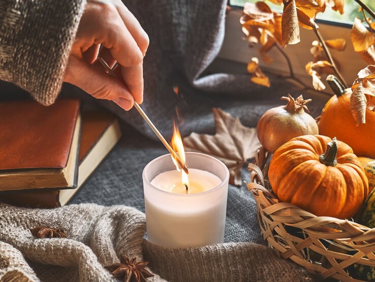 Herbstdeko aussortieren: Kerzen | © Shutterstock/Caterina Trimarchi Eine Hand in Strickpullover entzündet mit einem Streichholz eine Kerze, daneben Kürbisse in einem Korb und Bücher auf grauem Hintergrund. | © Shutterstock/Caterina Trimarchi