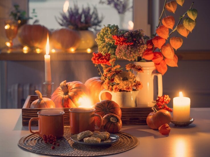Stimmungsvolles Arrangement mit Kerzenlicht, orangefarbenen Kürbissen, herbstlichem Blumenstrauß und zwei Tassen Tee auf weißem Tisch.