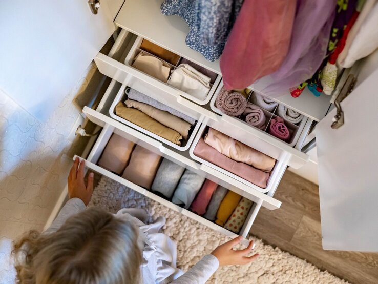 Ansicht von oben auf geöffnete Schubladen eines weißen Schranks mit ordentlich gefaltetem Stoff, ein Mädchen greift nach einer Schublade.