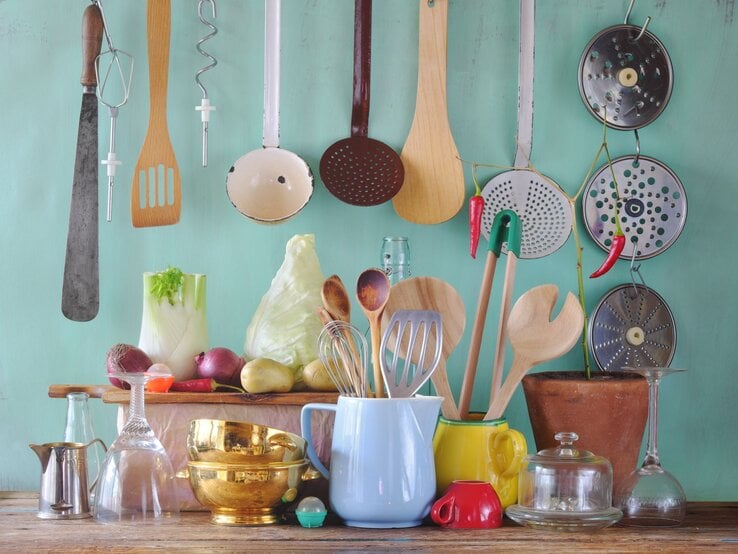 Küchenutensilien aus Holz, Glas und Metall sowie Gemüse wie Fenchel und Kohl arrangiert vor mintfarbenem Hintergrund auf einem Holztisch.