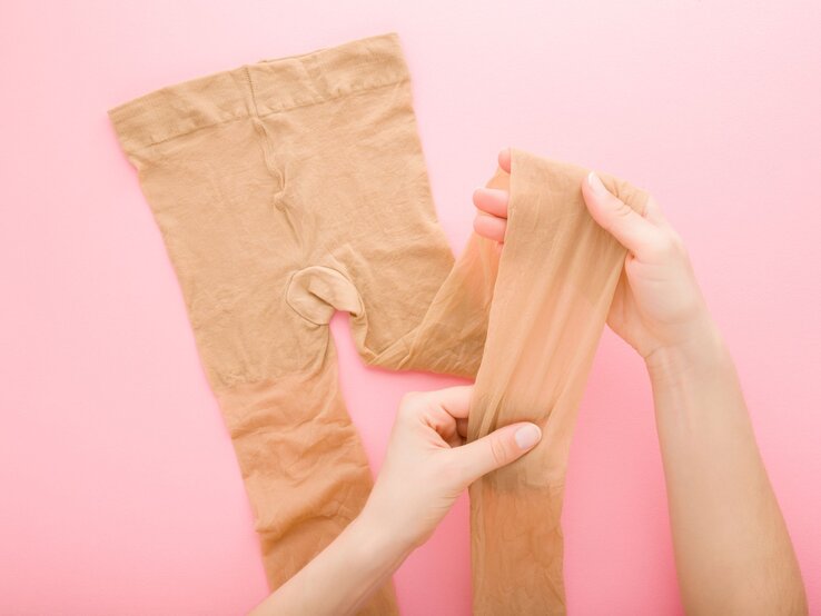 Nahaufnahme von Händen, die eine beige Strumpfhose auf einem rosa Hintergrund auseinanderziehen und prüfen. | © Shutterstock/FotoDuets