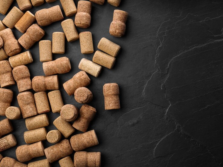 Viele unterschiedlich geformte Naturkorken liegen verstreut auf einer strukturierten schwarzen Steinfläche. | © Shutterstock/New Africa