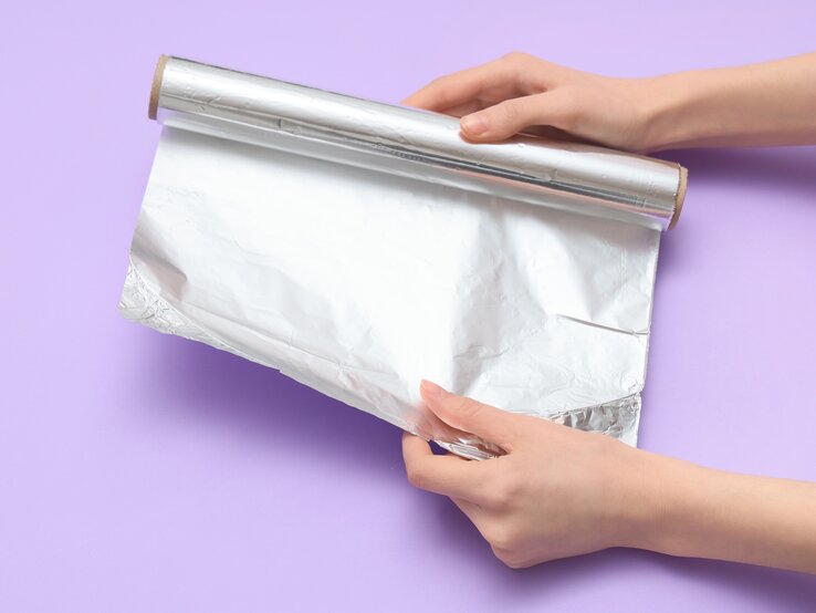 Eine Rolle Aluminiumfolie wird von zwei Händen auf einer lila Fläche entfaltet und glatt gezogen. | © Shutterstock/Pixel-Shot