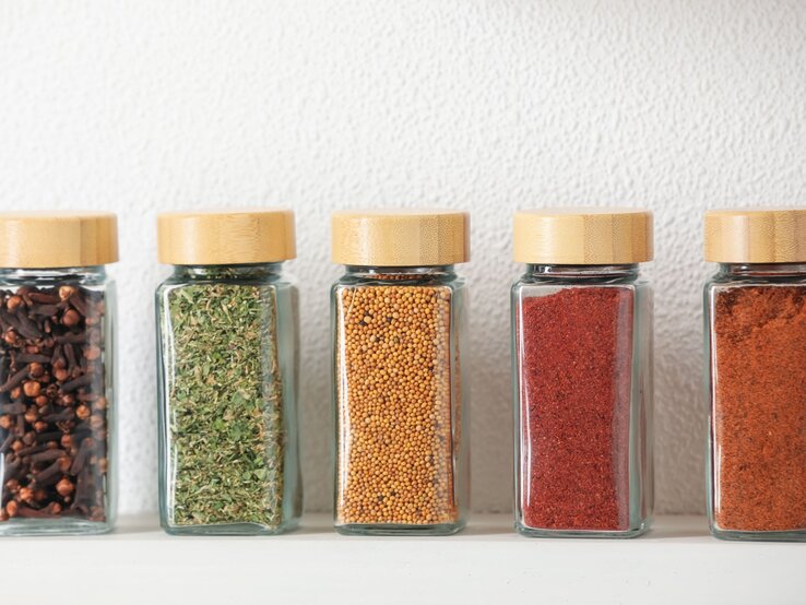 Fünf Gewürzgläser mit Bambusdeckeln, gefüllt mit Nelken, Kräutern, Senfkörnern und Gewürzpulvern, vor weißer Wand. | © Shutterstock/New Africa