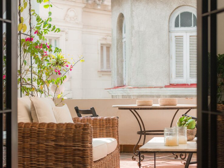 Eine sonnige Altbau-Loggia mit Korbsessel, weißen Polstern und kleinem Tisch, umrankt von pink blühender Bougainvillea.