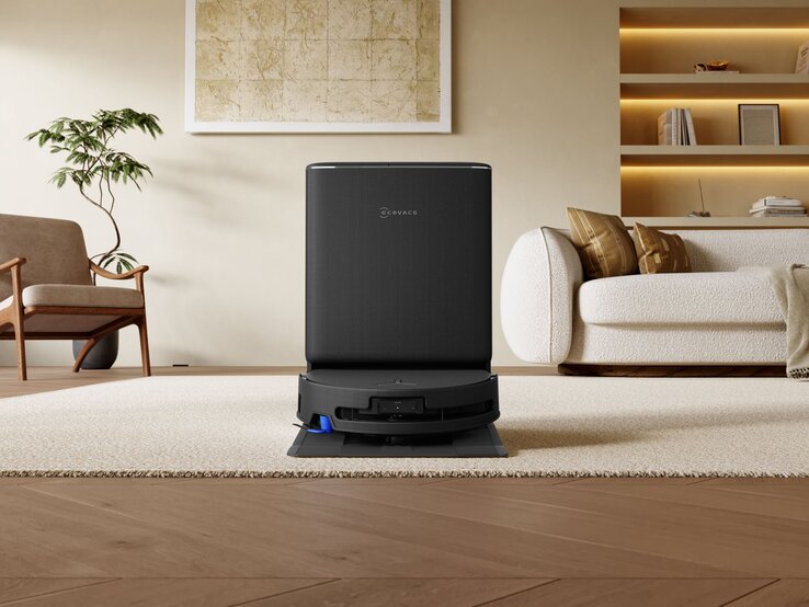 Schwarzer Staubsaugerroboter in Ladestation auf hellem Holzboden, daneben moderne Einrichtung mit Vase und Gemälde. | © ECOVACS