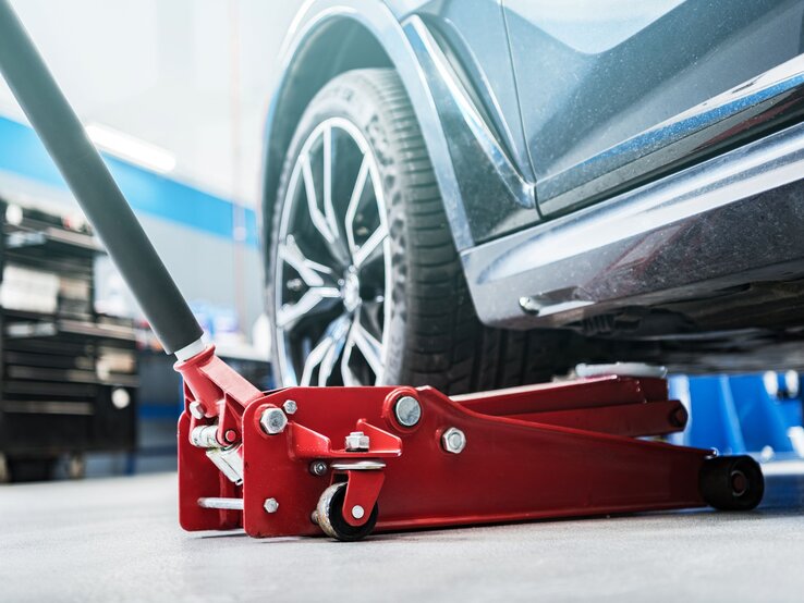 Ein roter Wagenheber hebt ein dunkles Auto in einer hellen Werkstatt an, das Vorderrad mit silberner Felge schwebt über dem Boden.