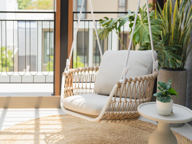   Heller Hängesessel mit beigem Kissen aus Seilgeflecht steht auf sonnigem Balkon neben Pflanzen und kleinem Beistelltisch.