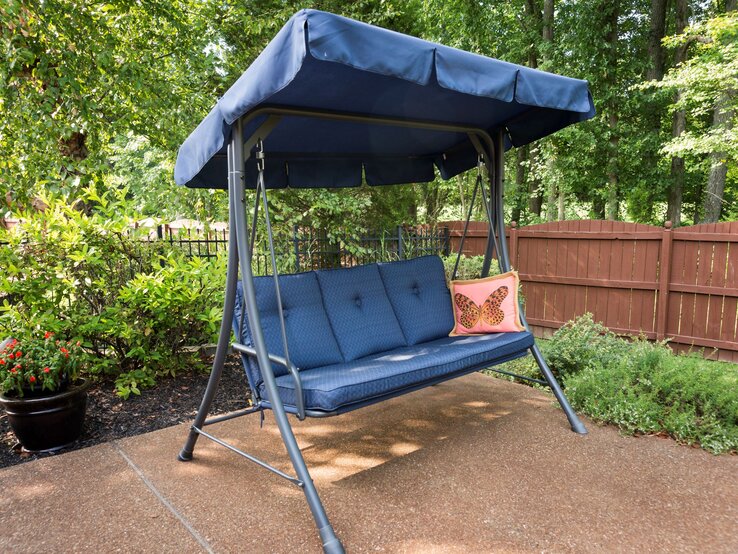   Dunkelblaue Gartenschaukel mit Sonnendach und Polstern steht auf Terrasse, umgeben von grünen Sträuchern und Holzzaun.