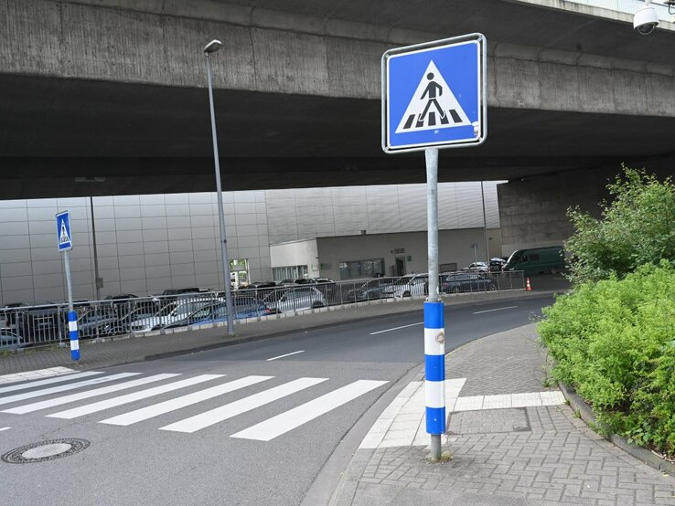 Ein Foto von einem städtischen Straßenbereich mit einem Fußgängerüberweg, erkennbar an den typischen weißen Streifen auf dem Asphalt. Direkt vor dem Zebrastreifen steht ein blau-weißes Verkehrsschild, das auf den Fußgängerüberweg hinweist. Die Umgebung wirkt urban mit einem Betonbauwerk im Hintergrund, unter dem die Straße hindurchführt. Am Straßenrand befindet sich eine Reihe geparkter Autos und auf der rechten Seite ist eine kleine Grünfläche mit Büschen zu sehen