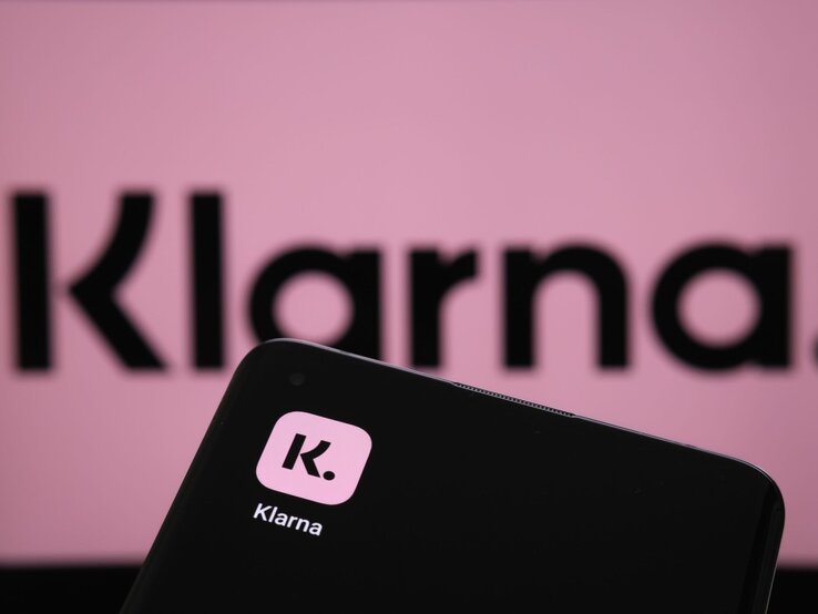 Im Vordergrund ist ein Smartphone mit dem Logo von Klarna, einer bekannten Zahlungs- und Shopping-App, zu sehen. Der Hintergrund zeigt unscharf das Klarna-Logo in Großbuchstaben auf einem rosa Hintergrund. Der Fokus liegt auf dem Smartphone