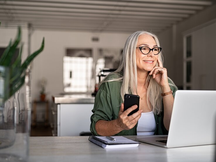 Dame höheren Alters sitzt vor dem PC mit dem Handy in der Hand