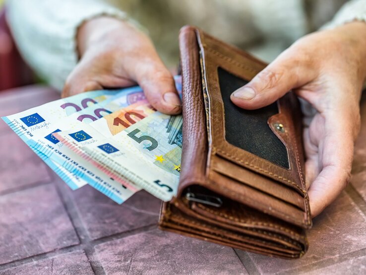 Eine Nahaufnahme von zwei Händen, die ein braunes Lederportemonnaie halten, aus dem mehrere Euro-Banknoten herausragen, darunter Scheine mit den Werten 5, 10, 20 und 50 Euro. Die Szene wirkt, als ob jemand gerade dabei ist, Geld zu zählen oder zu organisieren.