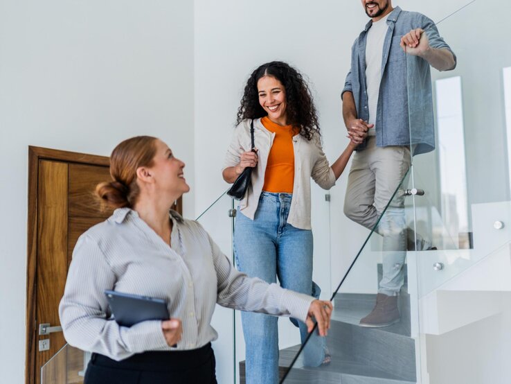 Eine Maklerin zeigt einer Frau und einem Mann eine Wohnung. Alle gehen hintereinander die Treppe runter. 