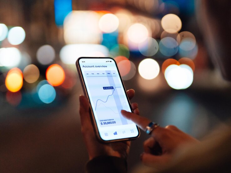 Frau guckt draußen auf ihr Smartphone. Darauf ist ein Aktienkurs zu sehen. 