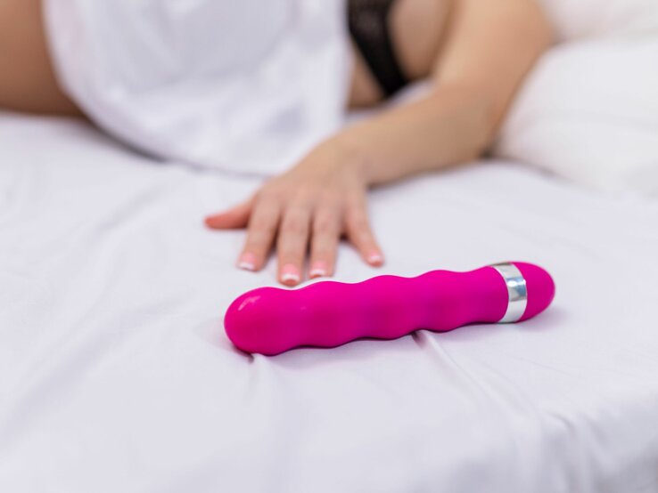 Ein pinkfarbener Vibrator liegt auf einem Bett, eine Frau liegt daneben, von der man nur den Arm sieht, der sich nach dem Sex Toy streckt