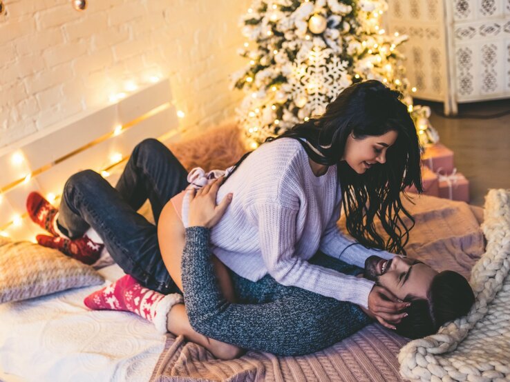 EIS Deluxe Adventskalender Ein Paar kuschelt verliebt auf einem Bett mit Lichterketten und Weihnachtsbaum im Hintergrund, beide wirken entspannt und fröhlich.