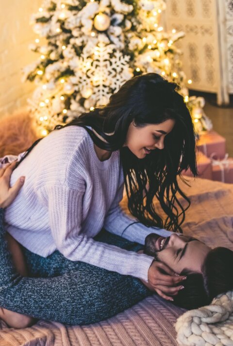 Ein Paar kuschelt verliebt auf einem Bett mit Lichterketten und Weihnachtsbaum im Hintergrund, beide wirken entspannt und fröhlich.