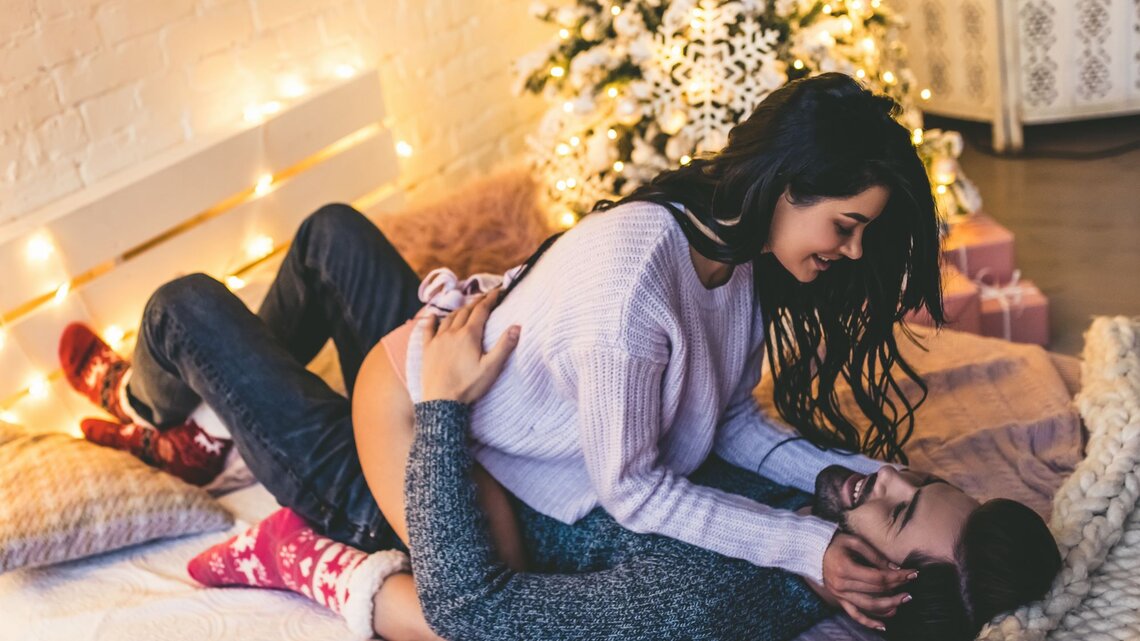 Ein Paar kuschelt verliebt auf einem Bett mit Lichterketten und Weihnachtsbaum im Hintergrund, beide wirken entspannt und fröhlich.