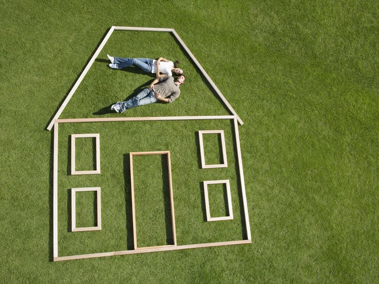 Junges Paar liegt träumend im Obergeschoss eines aus Holzrahmen geformten Hauses auf einer sattgrünen Wiese.