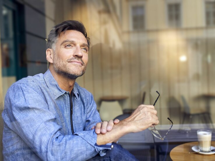 Ein gutaussehender Mann mittleren Alters sitzt an einem Bistrotisch im Freien mit einem Espresso, er schaut verträumt in die Ferne 