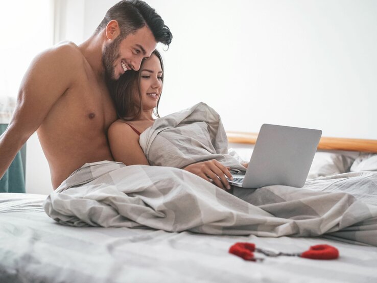 Auf dem Bild ist ein Paar zu sehen, das gemeinsam in einem Bett sitzt und ein Laptop betrachtet. Sie sind teilweise von Bettdecken bedeckt und scheinen Pornografie auf dem Laptop anzuschauen. Die Szene wirkt entspannt und vertraut, was auf eine Beziehung hindeutet.