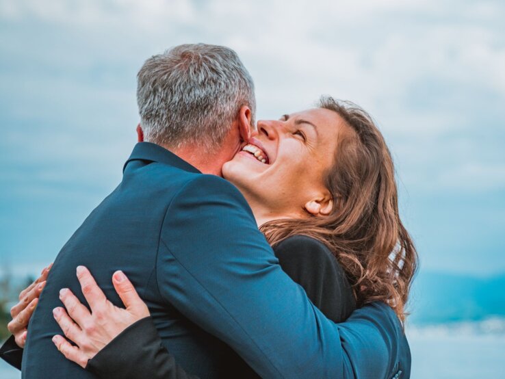Eine Frau mit braunen Haaren strahlt glücklich und umarmt einen Mann in blauem Anzug vor einem Seehintergrund.