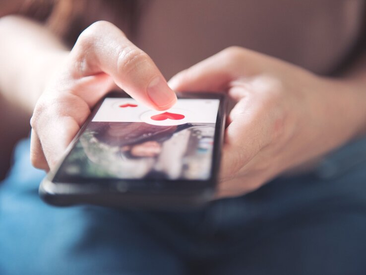 Mann hat Handy mit Dating-Portal in der Hand Nahaufnahme einer Hand, die ein Smartphone hält. Auf dem Bildschirm ist eine Dating-App geöffnet, die benutzende Person tippt auf ein Symbol in Form eines roten Herzens.