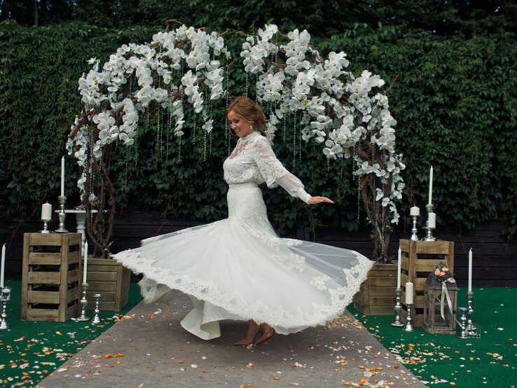 Braut tanzt vor Altar im Freien Eine lächelnde Braut mit hochgestecktem Haar dreht sich in einem weißen Spitzenkleid vor einem blumengeschmückten Hochzeitsbogen.
