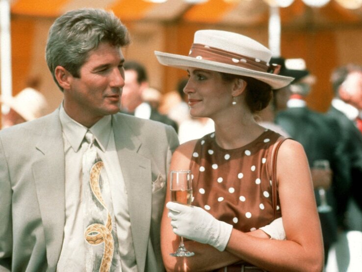 Pretty Woman Filmszene Eleganter Mann im hellen Anzug blickt lächelnd auf junge Frau im braunen Punktekleid mit Sektglas und weißem Hut bei Empfang.