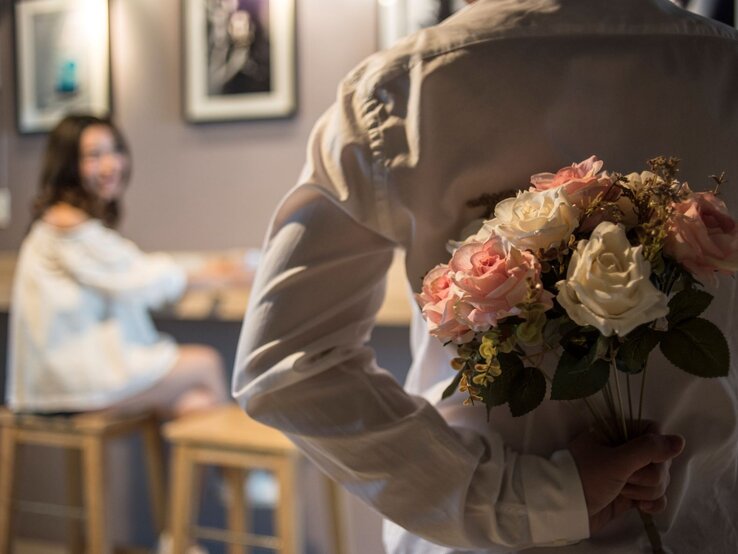 Ein Mann in weißem Hemd versteckt einen Rosenstrauß hinter dem Rücken, während eine junge Frau ihn lächelnd ansieht.