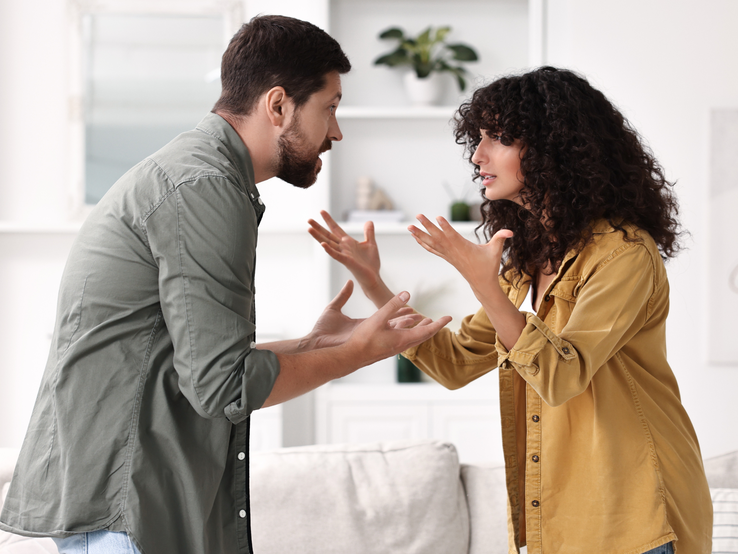 Ein junger Mann mit Bart und eine junge Frau mit Locken streiten gestikulierend im hellen Wohnzimmer mit Pflanze im Hintergrund.